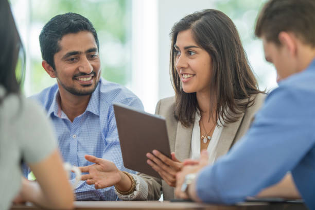 A multi-ethnic group of young business men and women in semi-casual office clothes are sharing ideas and holding a digital tablet in an indoor, sunlit office.